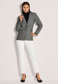 Grijze op maat gemaakte blazer met twee knopen, zwart coltruitje eronder, witte fitted broek met zijpiping en zwarte hakken met strikdetail.