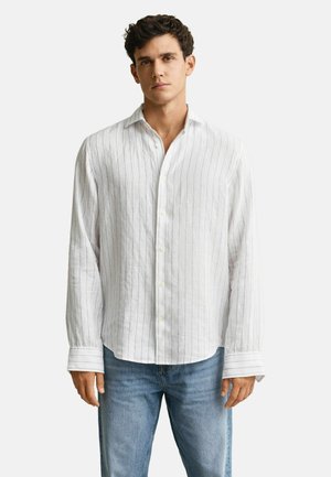 Hombre con camisa blanca de manga larga con botones y rayas verticales finas, y jeans azul claro, de pie frente a un fondo liso.