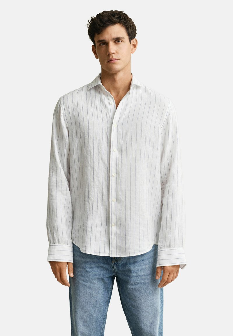 Homme portant une chemise blanche à manches longues avec de fines rayures verticales et un jean bleu clair, debout devant un fond uni.