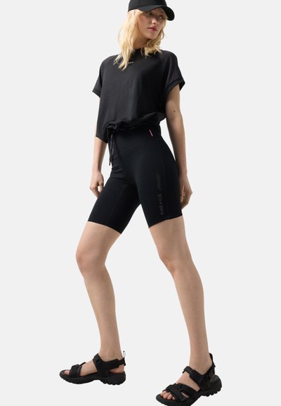 Femme portant une casquette noire, un t-shirt noir ample, un short de cyclisme noir ajusté avec le texte « FIRE + ICE » et des sandales sportives noires, debout avec une jambe en avant.