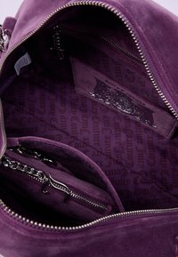 Interno di una borsa in suede viola con hardware argentato, dotata di una tasca con zip e logo impresso su una fodera a motivo con testo ripetuto.