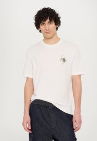 Camiseta de algodón blanca con un pequeño diseño bordado de una palmera en el lado izquierdo del pecho, mangas cortas y un cuello redondo.