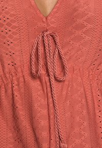 Korallivärinen paita, jossa on V-pääntie, teksturoitu timanttikuvio ja punottu nauha kaula-aukossa, jossa on reikäompeleet kauttaaltaan.