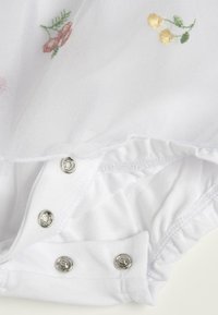 Un body blanc pour bébé avec des boutons-pression à l'entrejambe et de délicates fleurs roses et jaunes brodées sur un dessus en tissu transparent.