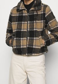 Chaqueta de forro polar con un patrón de cuadros en negro, marrón y gris, que cuenta con un cierre central de cremallera, bolsillos laterales y un cuello. Superficie texturada.