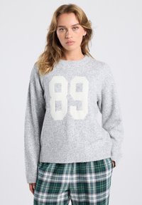 Femme aux cheveux châtain clair portant un pull gris avec un "89" blanc et un pantalon à carreaux verts, se tenant devant un fond uni.