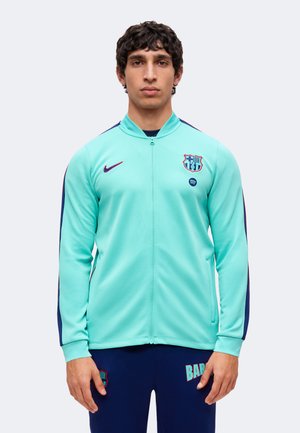 Jeune homme portant une veste Barcelona bleu clair avec une fermeture éclair et un pantalon bleu foncé, debout devant un fond clair uni.