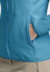 Veste ajustée en bleu clair avec une surface texturée, fermeture éclair et une poche latérale. Équipée de poignets élastiques et d'un cordon de serrage pour un ajustement.