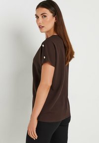Maglietta marrone a maniche corte con scollatura tonda e texture morbida. Presenta bottoni bianchi decorativi sulle spalle, progettata per un fit rilassato.