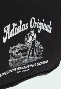 Čierna tkanina so bielou ilustráciou osoby pri pracovnom stole, obklopenej topánkami, s textom "Adidas Originals" nad ňou.