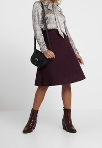 Svart axelremsväska, metallic ormtryckt blus med rosett, bordeauxröd utsvängd kjol och glänsande, bruna ankelhöga klackstövlar.