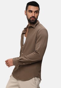 Camicia a maniche lunghe con bottoni in tessuto marrone testurizzato, con colletto e orlo curvo; abbinata a una canottiera bianca e pantaloni chiari.