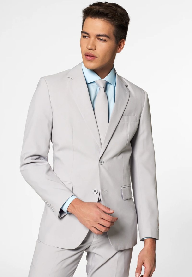 Homme en costume gris clair à deux boutons, chemise bleu clair et cravate assortie, debout, ajustant un bouton de veste d'une main.