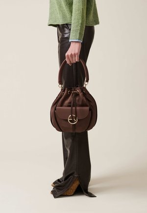 Brown textured leather handbag with a rounded shape, a top handle, a drawstring closure, and a front pocket with a circular gold accent.