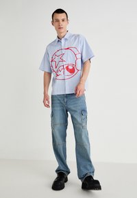Chemise à rayures bleues à manches courtes avec un grand motif graphique rouge, associée à un pantalon cargo léger bleu clair et des chaussures noires épaisses.
