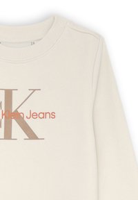 Felpa beige chiaro con collo a costine, caratterizzata da una grande "K" marrone e "Klein Jeans" in morbido testo pesca, tessuto texturizzato.