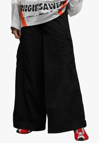 Pantaloni cargo neri a gamba larga con vestibilità rilassata, dotati di tasche laterali e dettagli di cuciture discrete. Abbinati a sneakers rosse e argento.