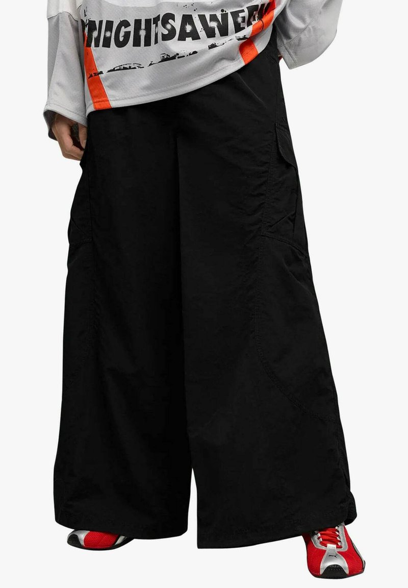 Pantaloni cargo neri a gamba larga con vestibilità rilassata, dotati di tasche laterali e dettagli di cuciture discrete. Abbinati a sneakers rosse e argento.