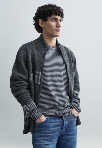 Grijze cardigan met een geribde kraag en manchetten, gedragen over een gestreept shirt met lange mouwen. Casual blauwe spijkerbroek maakt de outfit compleet.