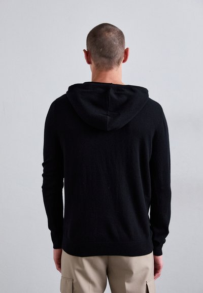 Svart hettegensere med en myk, strukturert finish, lange ermer og hette med snøringer, båret over lyse beige cargo-bukser.