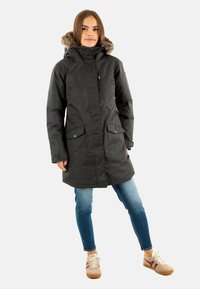 Parka noire avec une capuche en fausse fourrure, dotée d'une fermeture éclair sur le devant, de deux grandes poches avant et d'une coupe droite, associée à un jean bleu clair.
