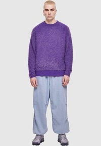 Pull en peluche violet avec un décolleté arrondi, des manches longues et une bande unie au bas. Associé à un pantalon ample bleu clair et des baskets.