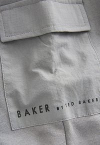 Harmaa kangas, jossa on teksturoidun ruudukon kuvio. Tunniste, jossa lukee "BAKER" suurilla mustilla kirjaimilla ja "by TED BAKER" pienemmällä fontilla alapuolella.