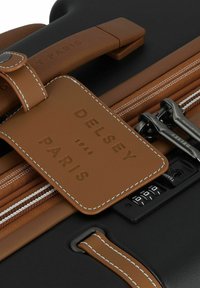 Valise noire avec une étiquette en cuir marron embossée, des accents de fermeture éclair et un cadenas à combinaison pour la sécurité.