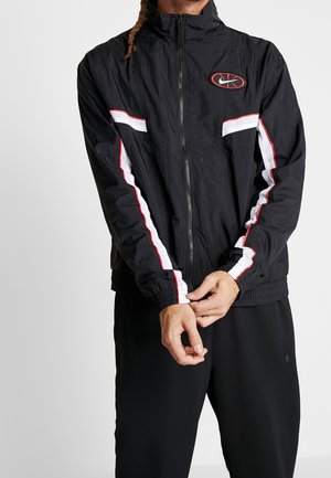 Homme portant une veste noire zippée intégralement avec des bandes blanches et rouges sur les manches et la poitrine, ajustant le poignet des deux mains.