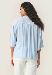 Blusa azzurro chiaro a maniche corte con vestibilità rilassata, caratterizzata da un dettaglio plissettato sul retro. Il tessuto sembra morbido e testurizzato.