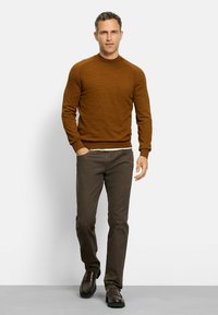 Brauner Strickpullover mit Rundhalsausschnitt, kombiniert mit dunkelbraunen Hosen und dunklen Schuhen. Der Pullover hat eine strukturierte Detailverarbeitung an den Schultern.