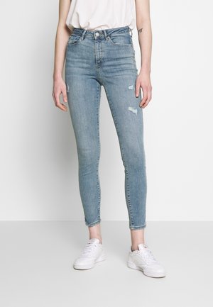 Personne portant un jean skinny bleu clair avec de petites déchirures et des baskets blanches, debout devant un fond uni.