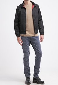 Svart bomberjacka, röd rutigt foder, beige tröja, grå jeans, svarta snörboots, stående figur, enfärgad bakgrund.