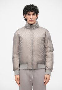 Jachetă bomber gri cu o textură matlasată și un model subtil cu logo. Dispune de guler în picioare, închidere cu fermoar și mansete ribbed.