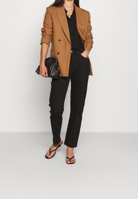 Blazer marron croisé, chemise noire, pantalon noir taillé sur mesure, pochette noire et tongs noires. Lignes épurées et couleurs neutres.