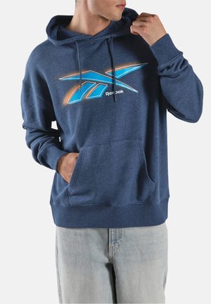 Mann trägt einen blauen Reebok-Hoodie mit großem blau-orangem Logo, hellen Jeans, Hand in der Tasche und zieht an der Kapuzenkordel.