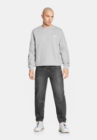 Szary sweatshirt z okrągłym dekoltem, ściągaczami przy rękawach oraz nadrukowanym logo na piersi, w zestawie z ciemnoszarymi dżinsami i białymi butami sportowymi.