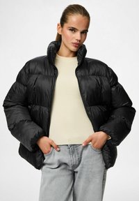Chaqueta de plumas negra con cuello alto, textura brillante y forma oversized, combinada con un suéter de punto beige claro y jeans grises.
