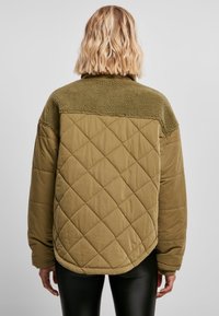 Oliven grønn quiltet jakke med teksturert fleecekrage, med avrundet hem og lange ermer. Bakfra viser den en flat, jevn finish.