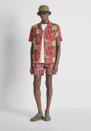 Homme portant un chapeau bob vert, des lunettes de soleil noires, une chemise à manches courtes verte et rouge à motifs ainsi qu'un short assorti, un t-shirt blanc et des chaussures vertes sans lacets.