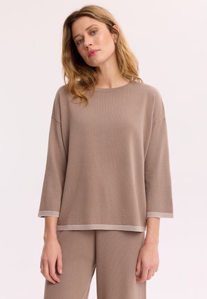 Kobieta z blond włosami do ramion, ubrana w luźną taupe długą bluzkę i pasujące spodnie na jednolitym tle.