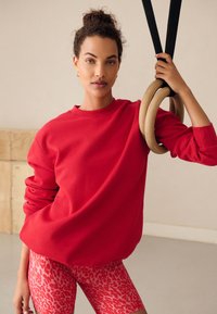 Röd sweatshirt med rund hals och långa ärmar, kombinerad med rosa leggings med djurprint, håller i trägymringar.