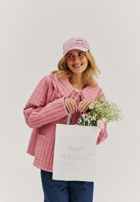 Růžová prošívaná bunda s velkým límcem, doplněná o růžovou čepici. Drží bílou tašku s logem "NAOKO" a kytici květin.