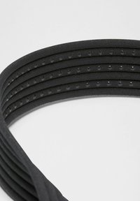 Tuvumā redzama melna elastīga josta ar četriem slāņotiem siksnām, katras siksnas malās ir mazi izvirzīti punkti, uz baltas virsmas.