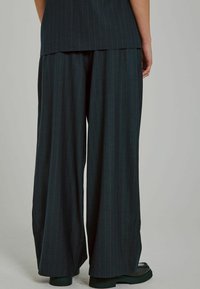 Pantaloni a gamba larga in tessuto verde scuro con righe verticali, caratterizzati da una texture liscia e una vestibilità rilassata, abbinati a scarpe nere massicce.
