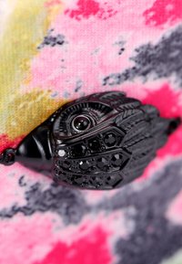Chiusura decorativa nera con dettagli intricati e accenti di gioielli, posizionata su uno sfondo di tessuto colorato e texture in rosa, giallo e grigio.