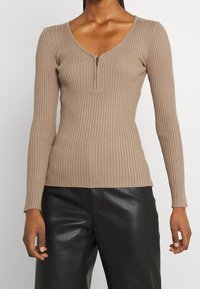 Beige ribbed long-sleeve top with a V-neck and quarter zip closure. Fitted design with texture details and a straight hem, paired with black pants.