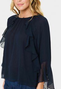 Marineblus i lätt tyg, med volangdetaljer längs halslinningen och ärmarna, avslappnad passform och genomskinlig textur.