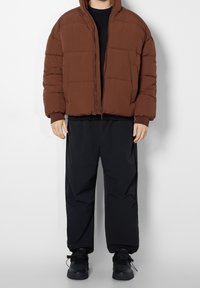 Giacca a vento marrone con cappuccio, caratterizzata da chiusura con zip e grandi pannelli cuciti, abbinata a pantaloni neri larghi e scarpe da ginnastica nere.
