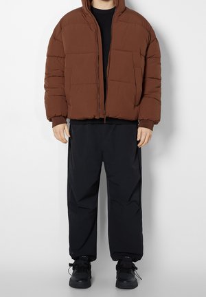 Doudoune marron avec capuche, dotée d'une fermeture éclair à l'avant et de grands panneaux cousus, associée à un pantalon noir ample et des baskets noires.